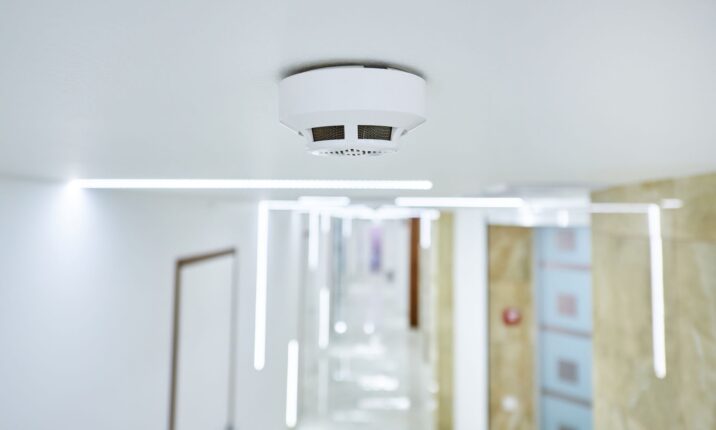 L'immagine ritrae un allarme antincendio installato sul soffitto di un passaggio interno. L'apparecchio, di forma rotonda e colore bianco, risalta su uno sfondo chiaro. Il corridoio è rischiarato da illuminazione lineare a soffitto, conferendo un'atmosfera luminosa e contemporanea. In lontananza si scorgono porte e pareti, indicando che si tratta di un ambiente chiuso, probabilmente un edificio adibito a uffici o una struttura aperta al pubblico.